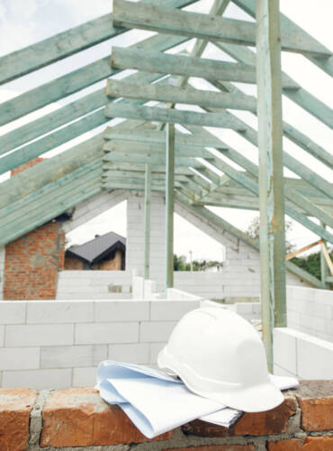 Architecture and House building concept. Hard hat with house blueprints on brick masonry on background of unfinished wooden roof framing. Safety helmet on construction site. Copy space
