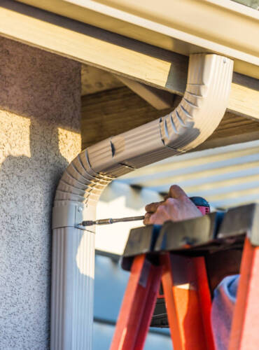 Worker Attaching Aluminum Rain Gutter and Down Spout to Fascia of House.