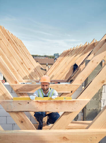 Serious builder in a hardhat kneeling near a wooden bar and using a level instrument
